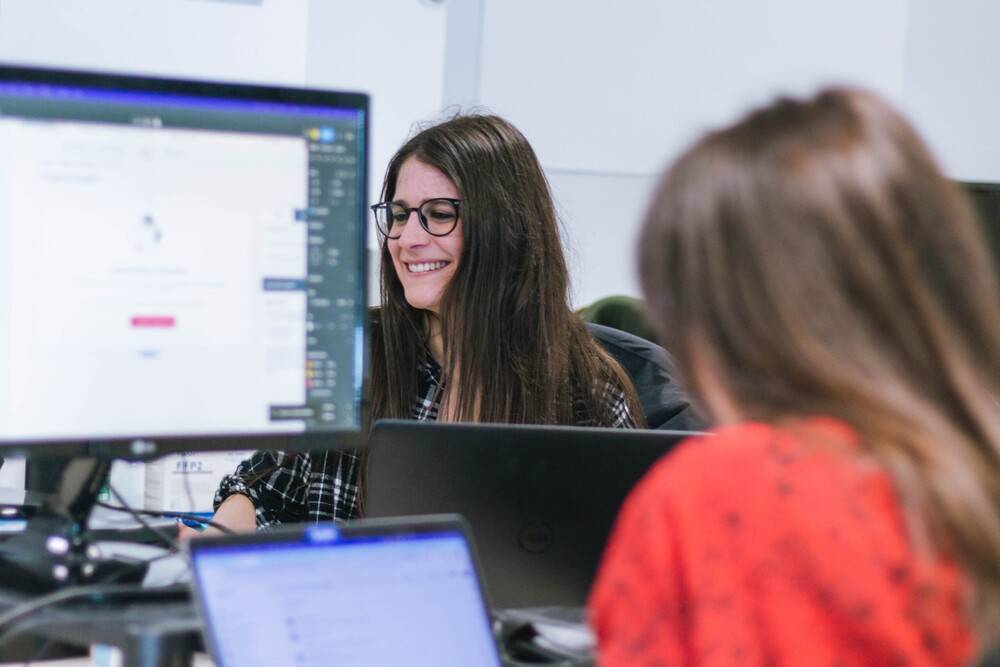 Una escena de oficina moderna en la que dos mujeres trabajan frente a sus ordenadores portátiles. En primer plano, desenfocada, se ve una persona de espaldas con una blusa roja, al fondo, en el centro de la composición, destaca una mujer con gafas y cabello oscuro, largo y suelto que sonríe mientras mira la pantalla de su portátil.