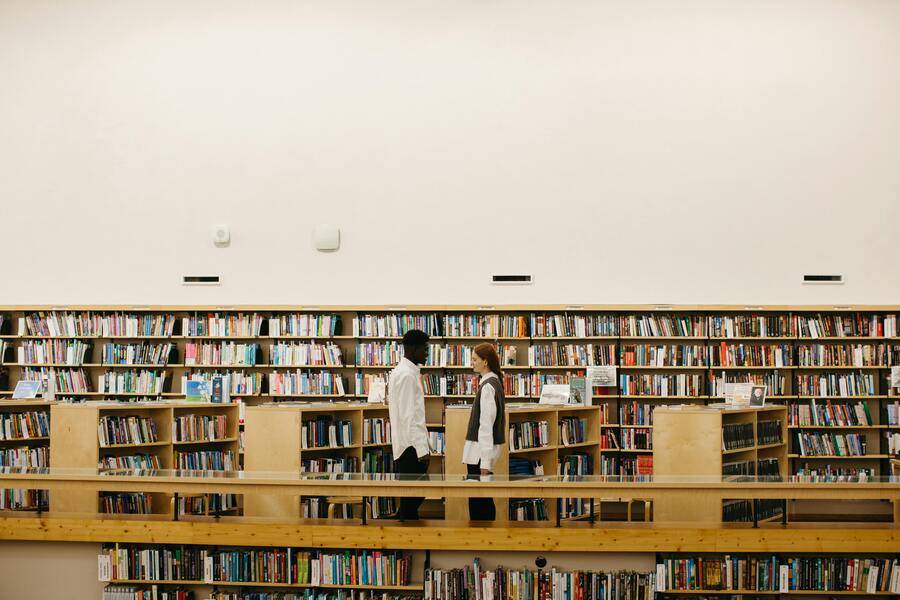 Innovación educativa: de sistemas legacy a arquitecturas inteligentes con SofIA y SOKAI 3 Dos estudiantes dialogando en una biblioteca rodeada de estanterías llenas de libros, reflejando la innovación educativa a través de la interacción colaborativa.