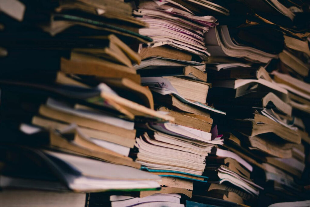 una gran pila desordenada de libros y cuadernos. Los tomos tienen las tapas gastadas, bordes doblados y papeles amontonados de forma caótica, lo que transmite sensación de acumulación y desorden.