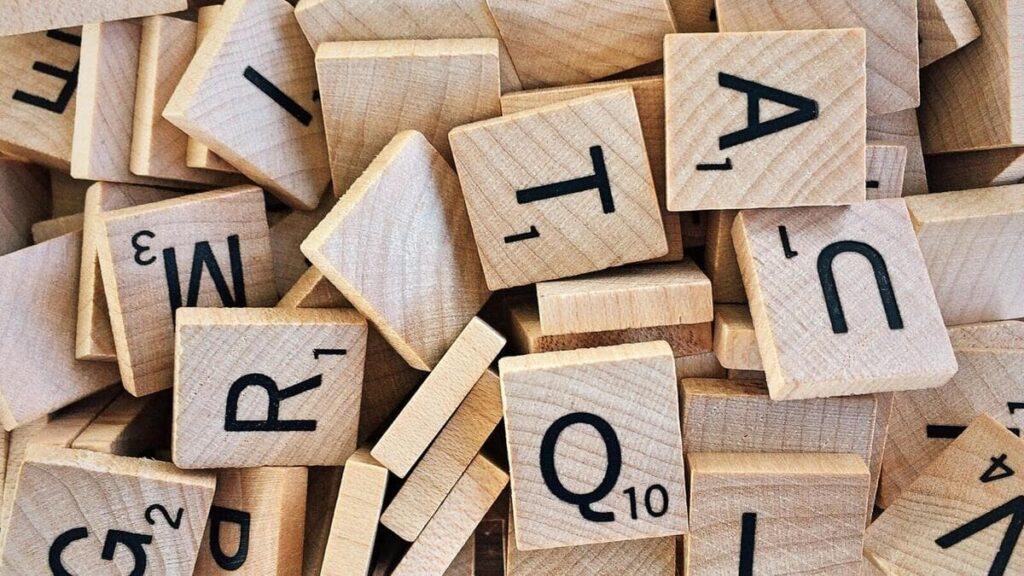 Piezas de madera con letras y números tallados, similares a las de Scrabble. Las piezas están amontonadas de forma desordenada, destacando letras como Q, R, M, T, A y U.