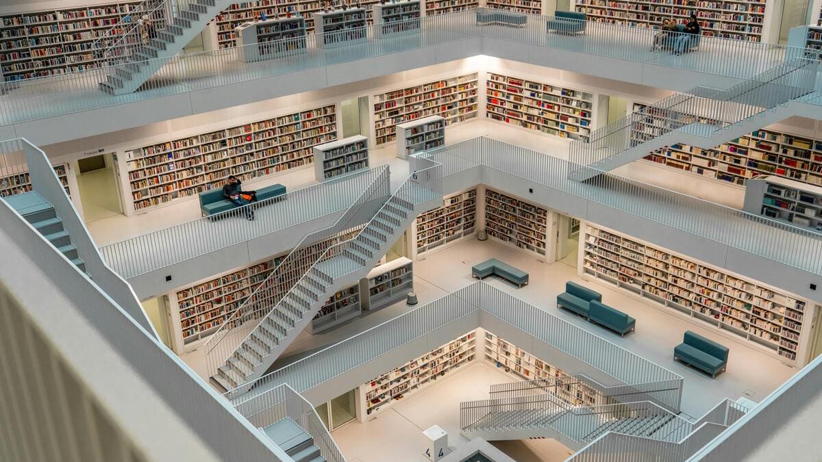Una biblioteca moderna y minimalista, caracterizada por su arquitectura abierta y geométrica. El espacio está organizado en varios niveles conectados por escaleras blancas, con estanterías repletas de libros que cubren las paredes de cada planta.