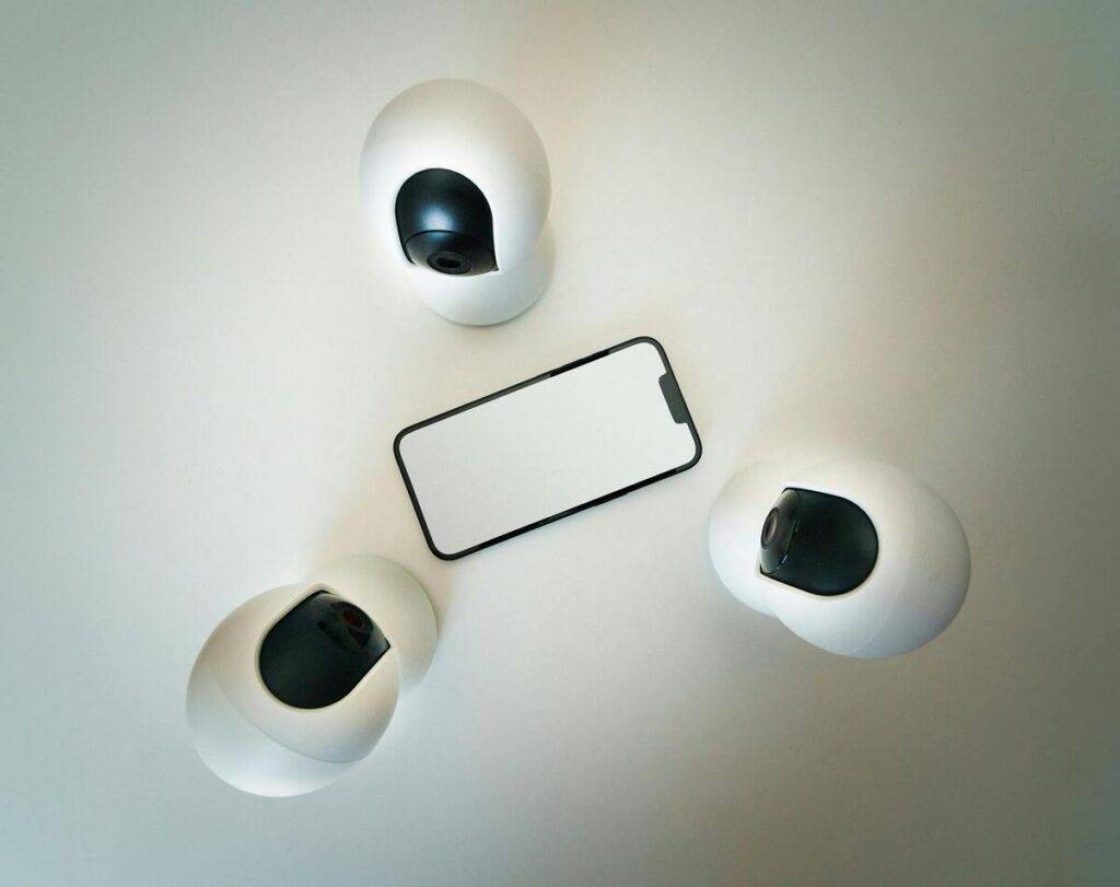 Tres cámaras de videovigilancia rodean un teléfono móvil sobre una mesa blanca. La composición simboliza la vigilancia digital en entornos de proctoring.