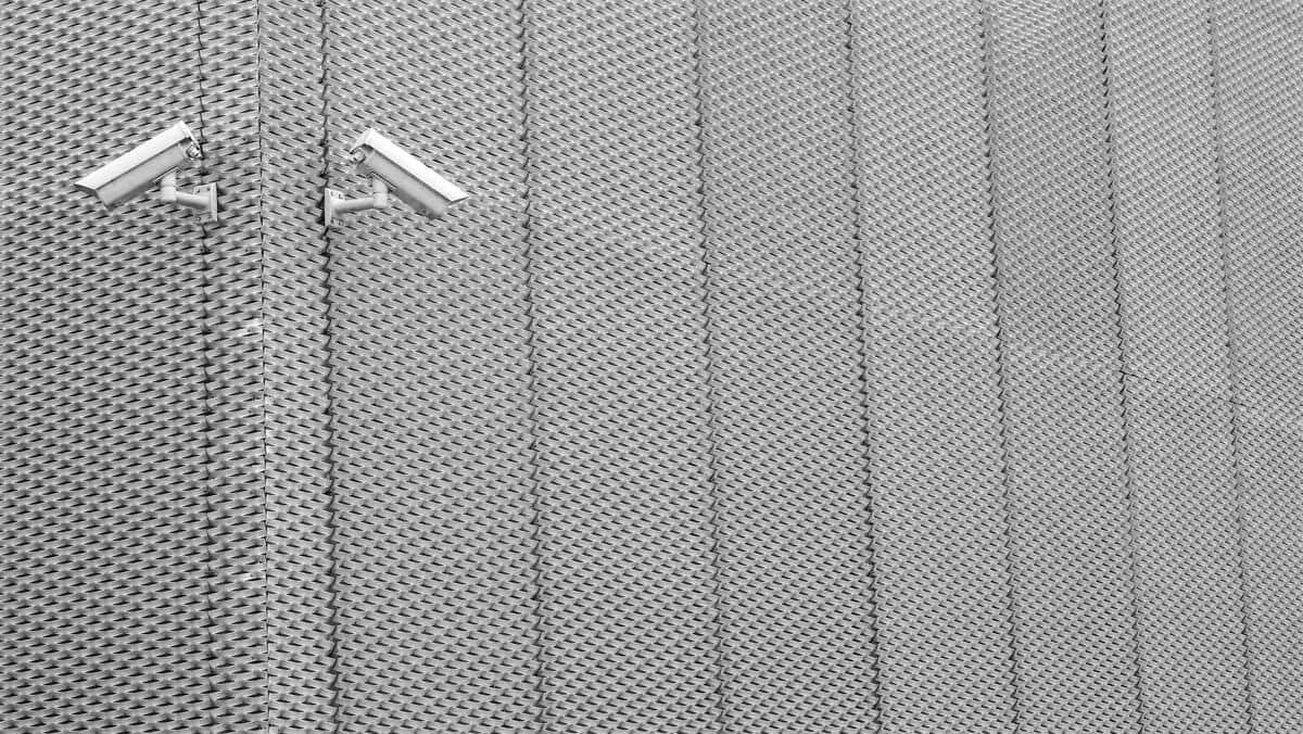 La fotografía muestra dos cámaras de seguridad instaladas en una pared metálica con un patrón geométrico repetitivo. Las cámaras están dispuestas de forma simétrica y orientadas en direcciones opuestas.