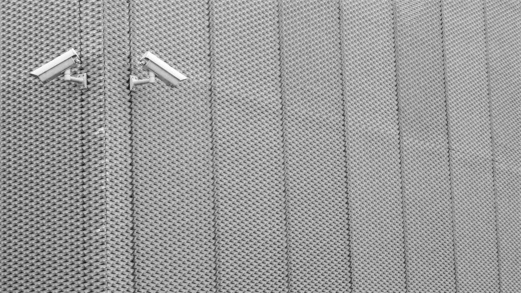 La fotografía muestra dos cámaras de seguridad instaladas en una pared metálica con un patrón geométrico repetitivo. Las cámaras están dispuestas de forma simétrica y orientadas en direcciones opuestas.