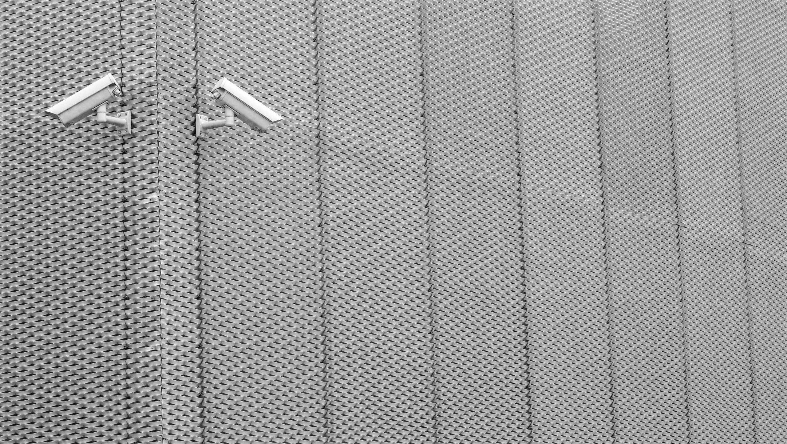 La fotografía muestra dos cámaras de seguridad instaladas en una pared metálica con un patrón geométrico repetitivo. Las cámaras están dispuestas de forma simétrica y orientadas en direcciones opuestas.