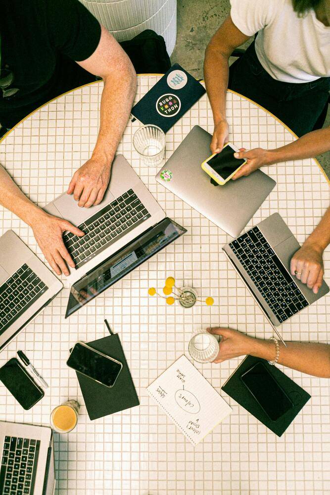 Personas trabajando en grupo alrededor de una mesa redonda, cada una con su portátil y teléfonos móviles. Se observan cuadernos, notas y bebidas, en un entorno colaborativo.