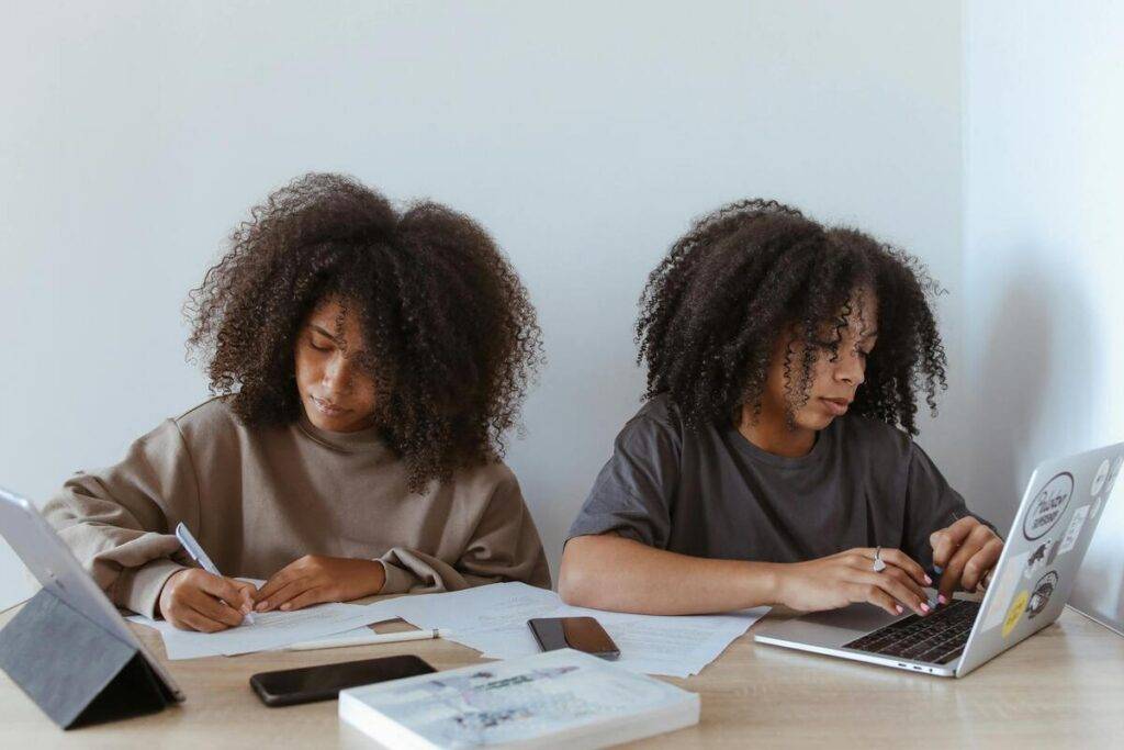 Dos jóvenes escribiendo y trabajando con dispositivos. Una toma notas en papel y la otra utiliza una laptop, ambas rodeadas de materiales de estudio.