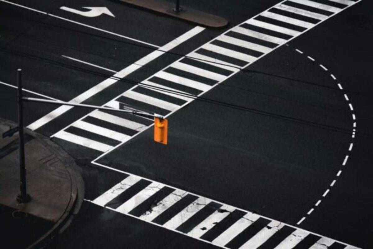 Una intersección urbana vista desde arriba, con marcas viales blancas claramente definidas sobre el asfalto oscuro. Se aprecia un semáforo amarillo en el centro de la escena, que contrasta con la geometría precisa de los pasos peatonales